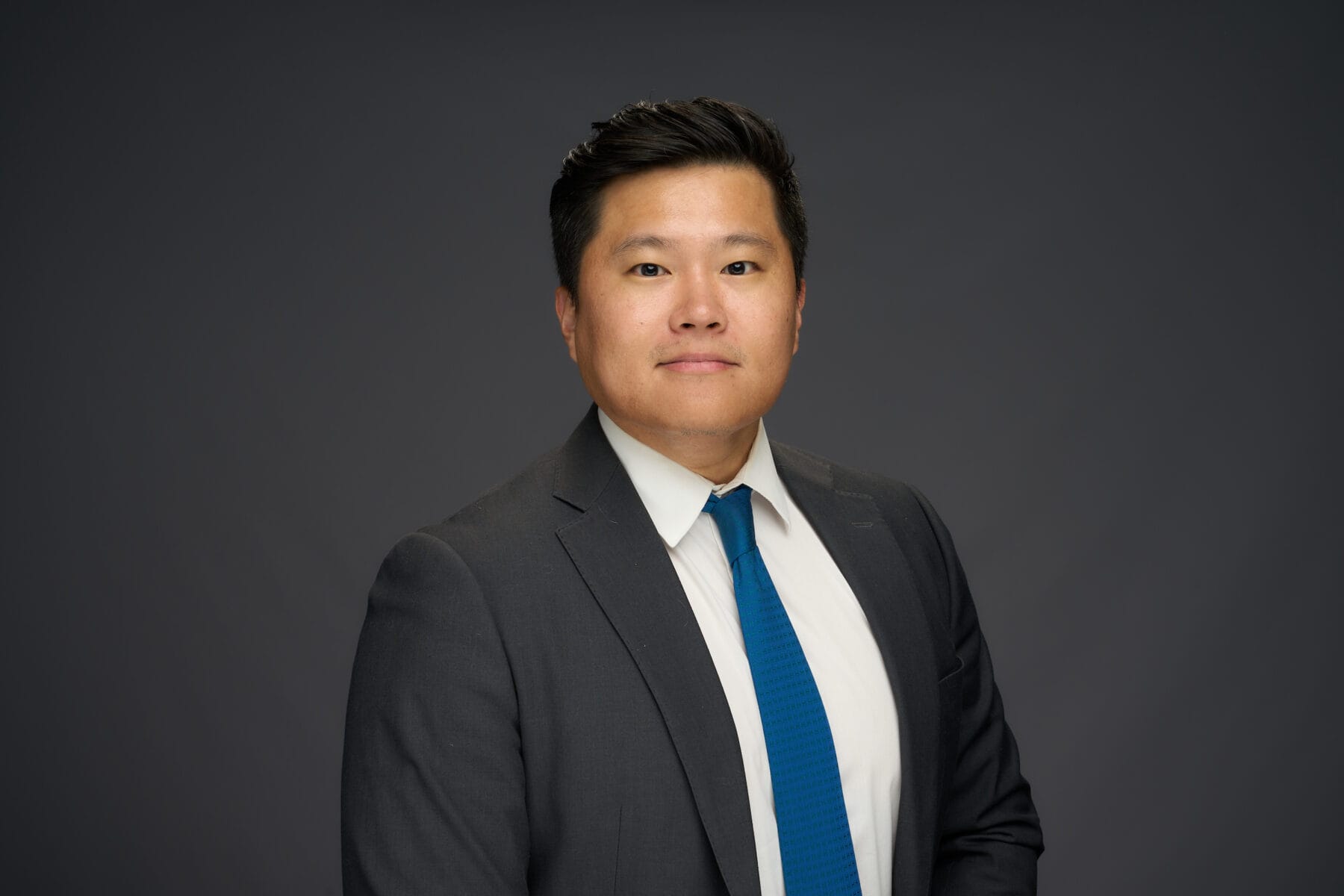 A man in a dark suit, white shirt, and blue tie poses for a professional studio portrait against a plain dark background.