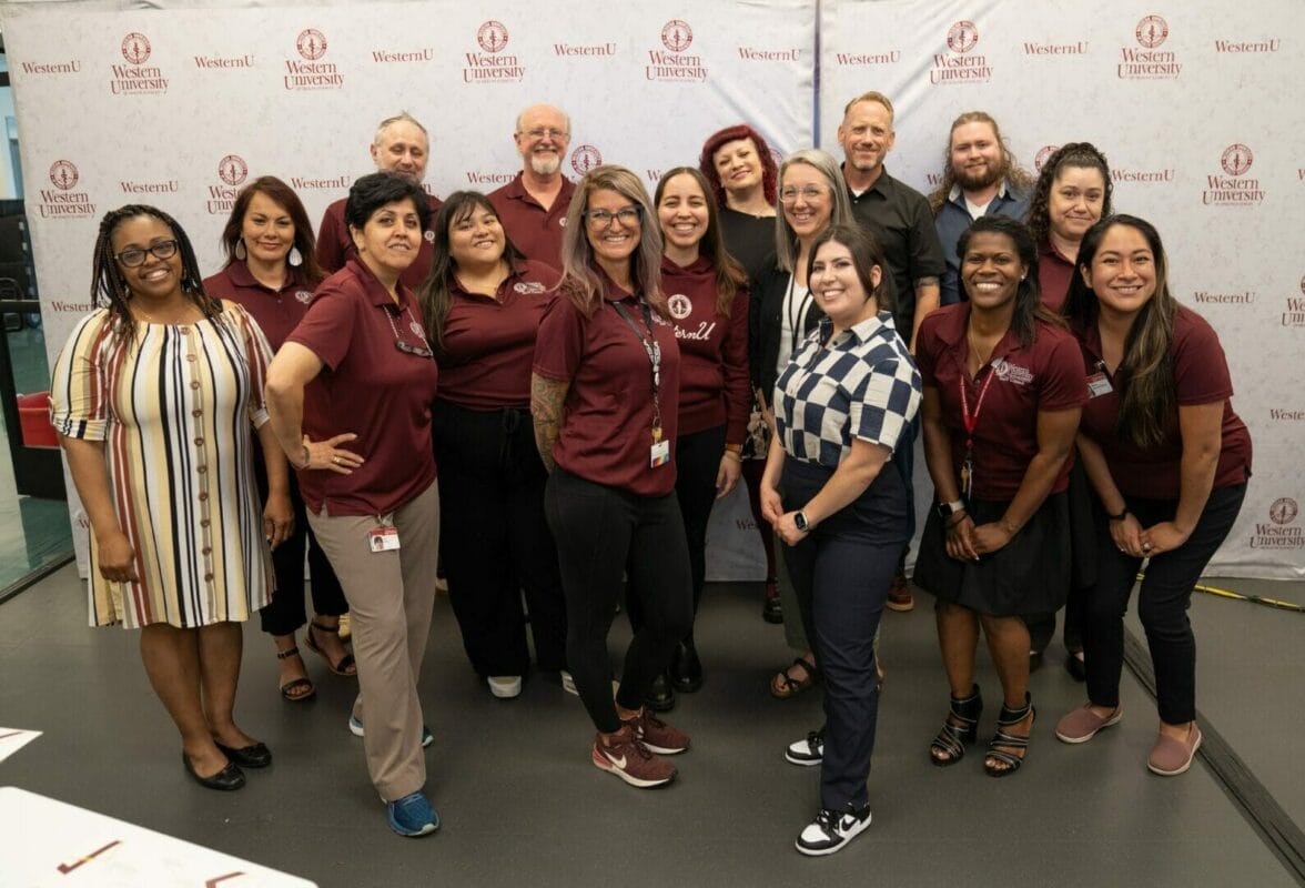 A group of people stand together for a photo in front of a backdrop with repeated logos, all smiling towards the camera. Some are wearing maroon shirts with a university logo.