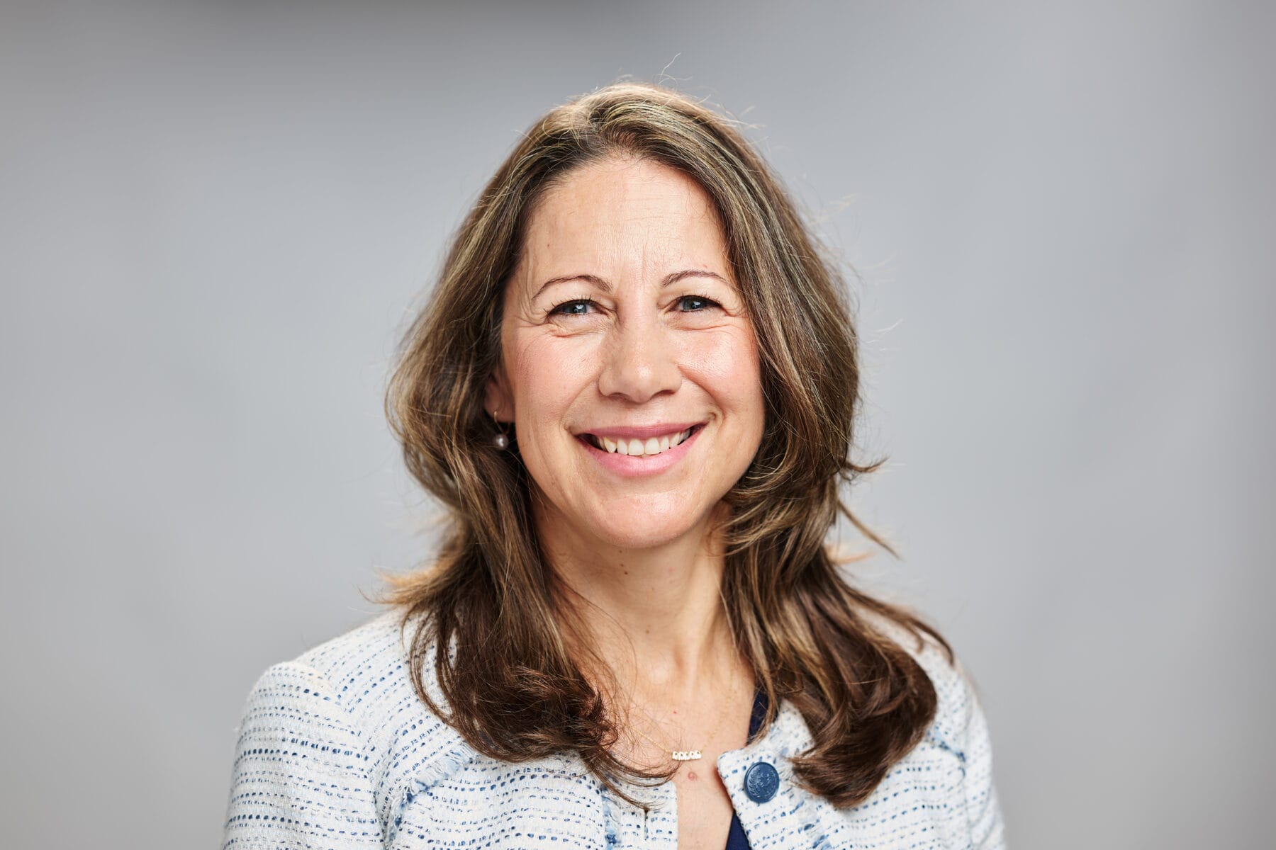 A woman with shoulder-length brown hair and a light blue blazer smiles in front of a plain gray background.