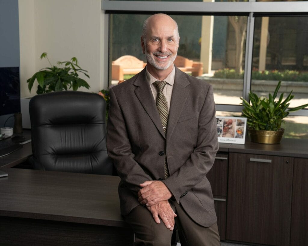 A smiling man in a brown suit and tie sits on the edge of a desk in a well-lit office with plants and a window in the background.