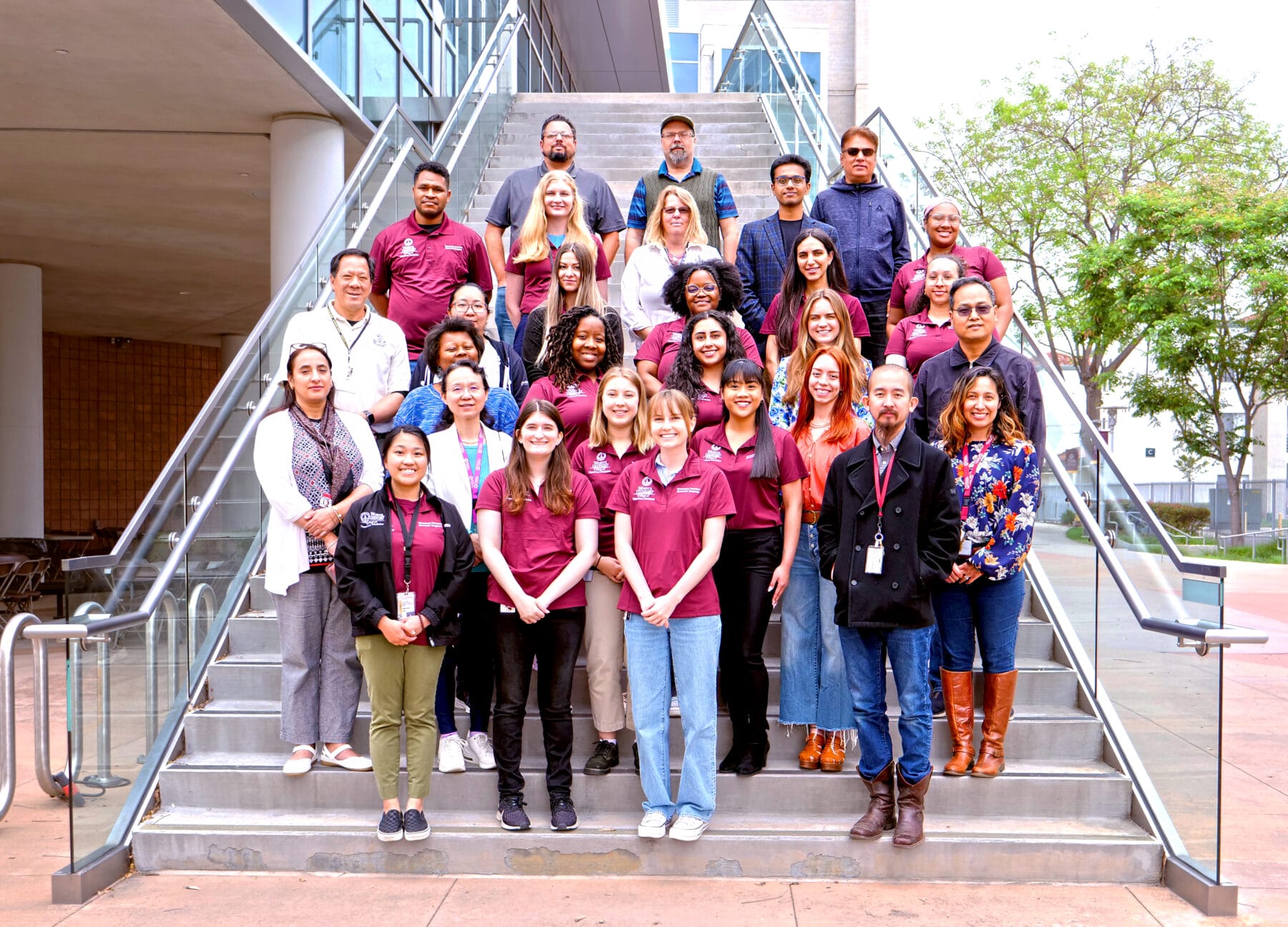 A group of people, some wearing maroon shirts, stands and sits on a series of steps outdoors in front of a building, posing for a group photo.