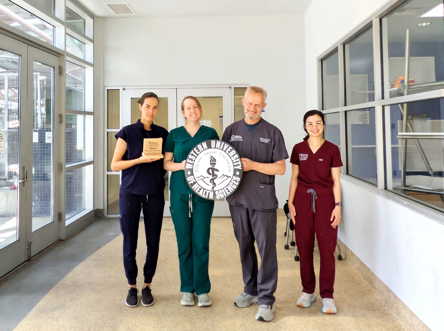 Four people in medical scrubs stand indoors, holding a Western University of Health Sciences sign and a plaque, posing for a group photo.