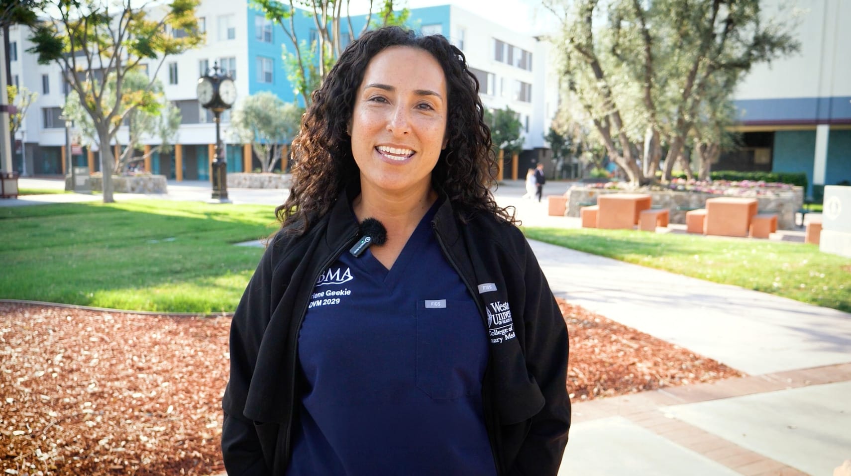 WesternU CVM student Darlene Geekie, who is wearing navy scrubs and a black jacket, stands outside on the WesternU California campus with buildings, trees, and a clock in the background.