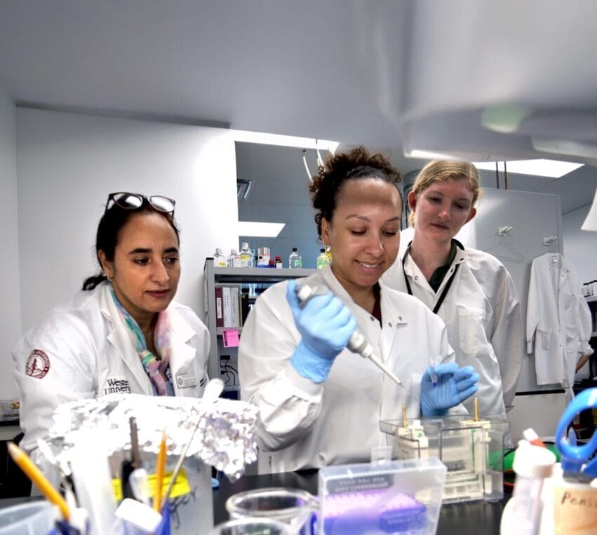 Three scientists in lab coats work together at a laboratory bench, using pipettes and various lab equipment in a research setting.