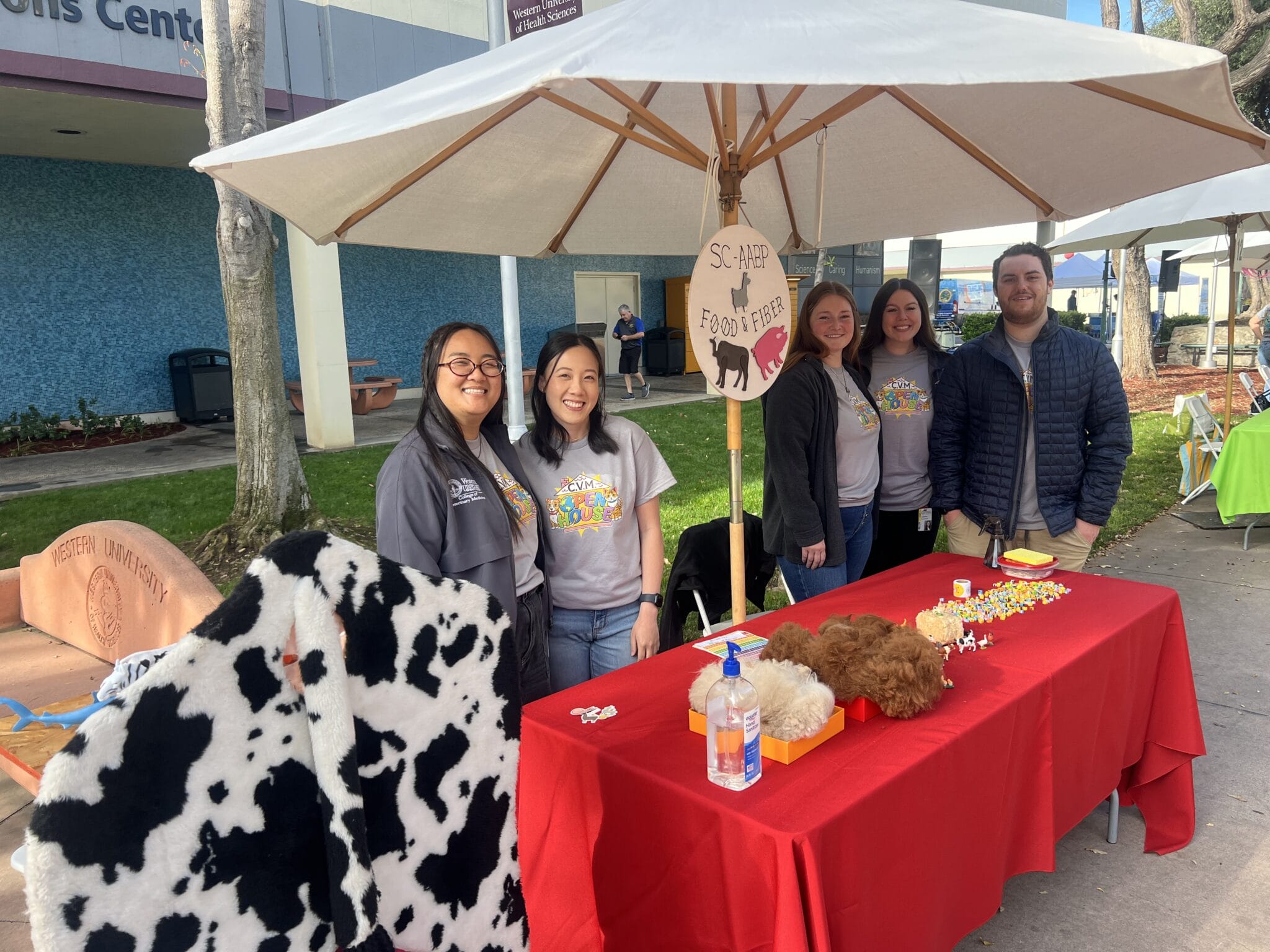 Five people stand behind a table with red cloth, animal props, and hand sanitizer under a white umbrella outdoors for a "Share a Furry Pet" event.