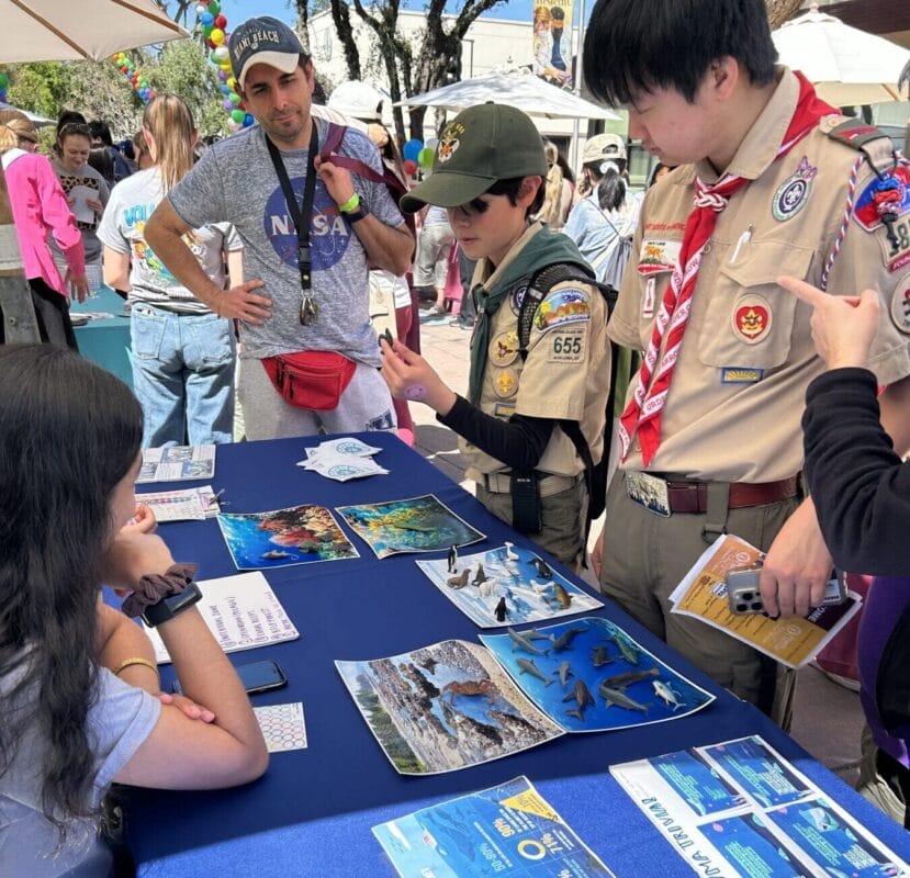 Two scouts in uniform talk to a seated visitor at an outdoor informational booth with brochures and posters about ocean wildlife on a blue tablecloth. Other people are seen in the background.