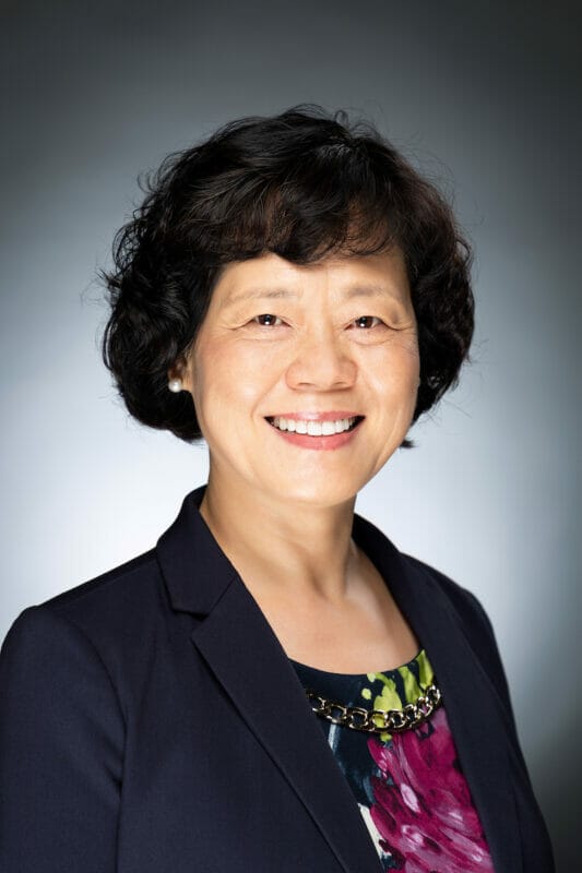 Woman with short curly hair wearing a dark blazer and floral top, smiling at the camera against a neutral background.